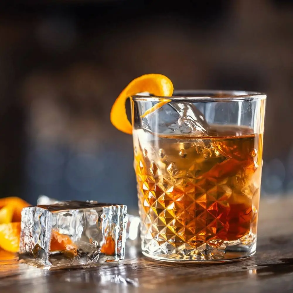 A old fashioned cocktail sitting on a wooden bar surrounded by ice and orange zest.